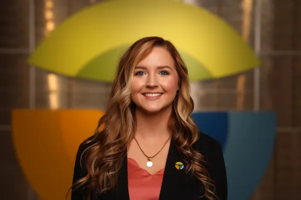 Smiling woman with long wavy hair wearing black blazer and coral top, colorful abstract background behind her