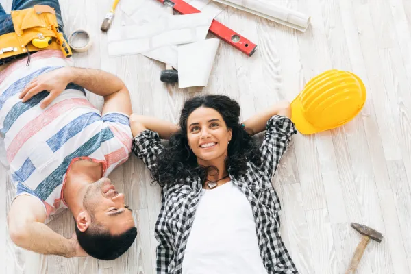 A couple of home renovators lying on the floor surrounded by tools and a yellow construction helmet.