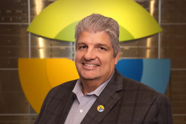 Smiling man in a gray suit with gray hair stands in front of a colorful yellow, green, and blue wall logo.