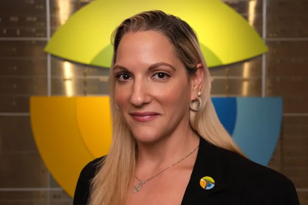 Professional woman with blonde hair wearing black blazer and colorful pin standing in front of geometric yellow and blue background.