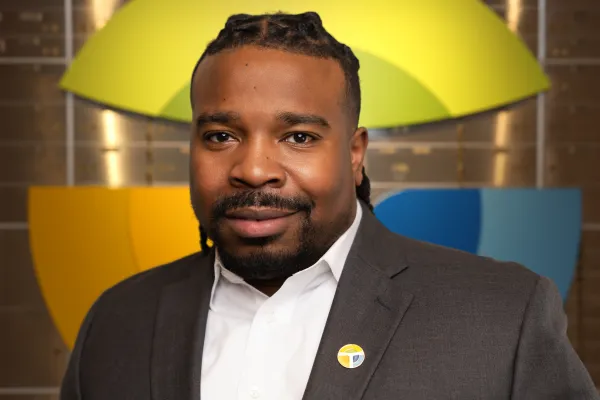 Professional man in suit with badge stands before colorful geometric logo background, smiling confidently.