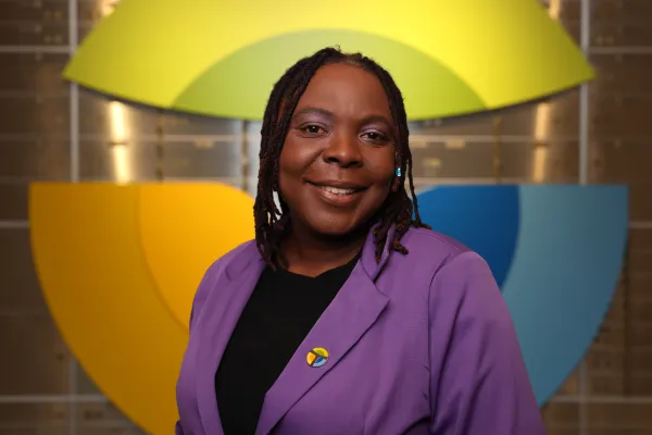 Smiling woman in purple blazer with colorful pin stands against a backdrop of overlapping yellow and blue shapes.