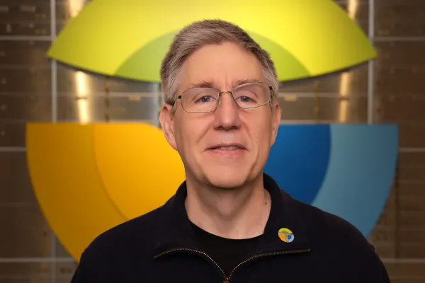Smiling middle-aged man with glasses wearing a dark jacket and colorful logo pin in front of large geometric backdrop.