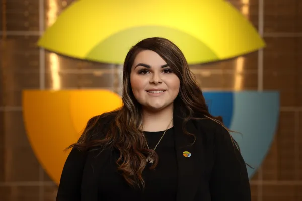 Smiling woman with long brown hair wearing a black blazer and colorful pin in front of a multicolored abstract background