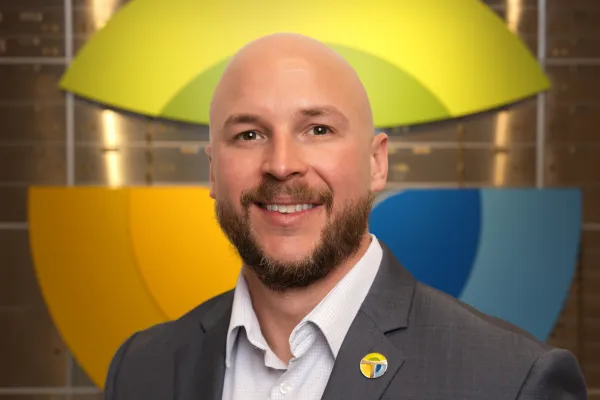 Smiling bald man with beard in gray suit and white shirt, standing in front of colorful abstract logo wall.