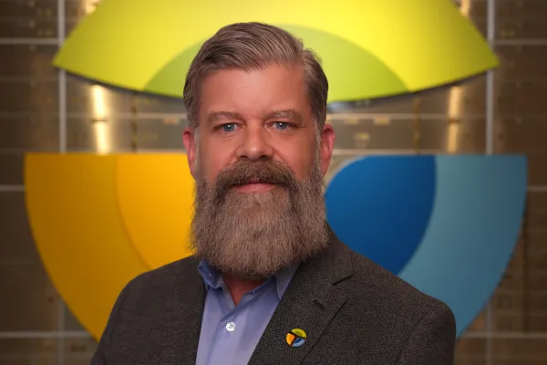 Man with a gray beard and suit jacket smiling in front of a colorful circular logo background indoors.