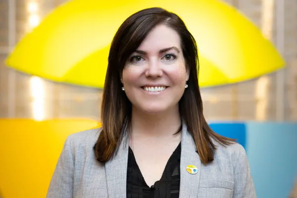 Smiling woman with brown hair wearing a gray blazer and pin, with colorful yellow and blue background.