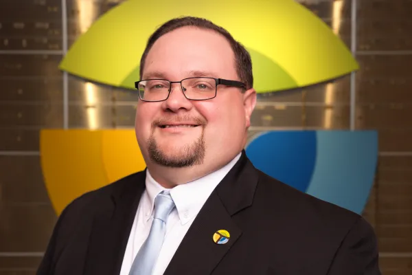 Smiling man wearing glasses and a suit with a light blue tie and pin standing in front of colorful logo backdrop.