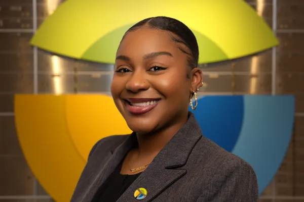 Smiling woman with natural hair wearing a navy cardigan and colorful pin against a geometric yellow and blue background.