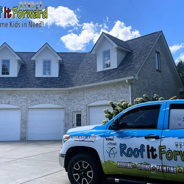 Blue and white Roof It Forward truck parked in driveway of large house with gray roof and garage doors