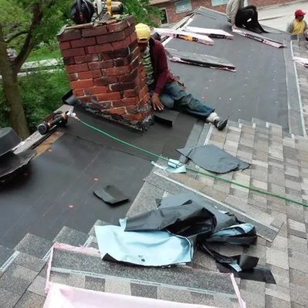 Roofers installing shingles and sealing a chimney on a residential roof under cloudy skies.