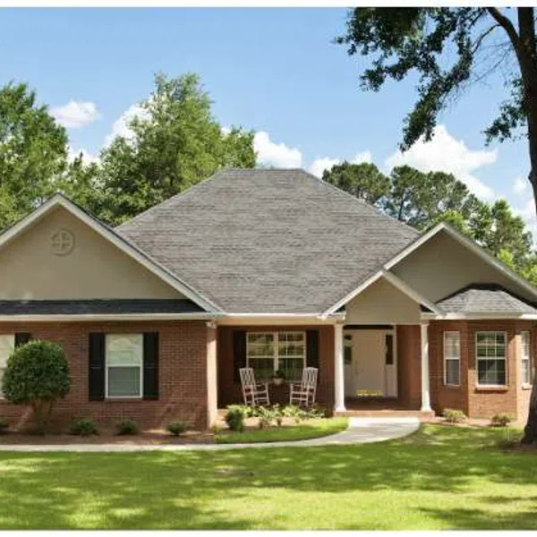 Single-story brick house with gray roof, front porch, lawn, and surrounding trees on a sunny day