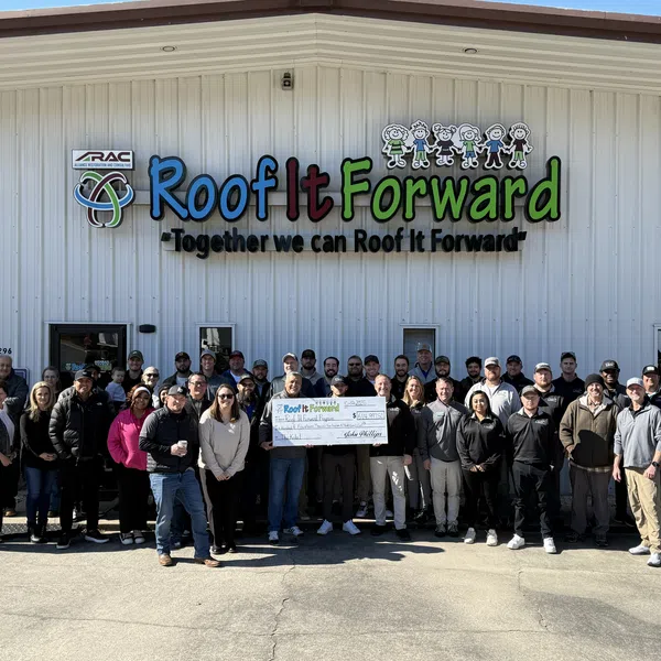 Group of Roof It Forward team members standing outside building holding a large check for a charity event.