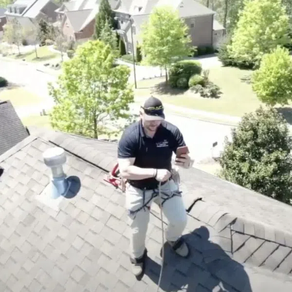 Man in safety harness standing on rooftop inspecting area with smartphone on sunny day in residential neighborhood.