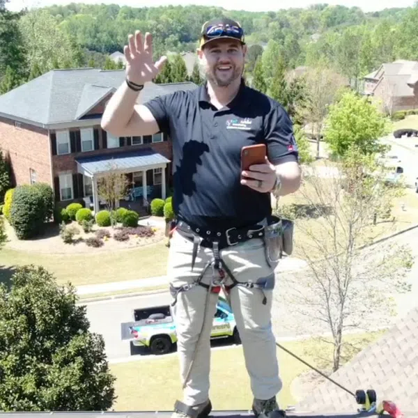 A smiling worker on a roof, waving and holding a phone, with houses and greenery in the background.