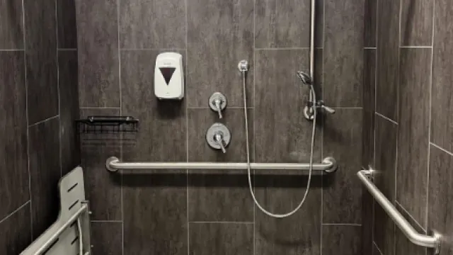 Accessible shower with folding seat, grab bars, hand shower, and soap dispenser in tiled bathroom.