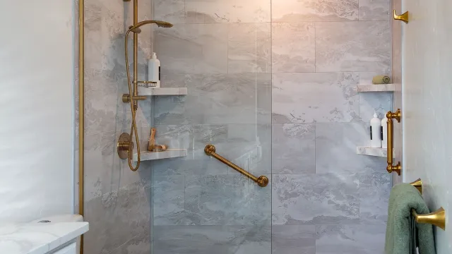 Modern bathroom with gold fixtures, pebble stone shower floor, gray tiles, glass shower door, and white vanity.