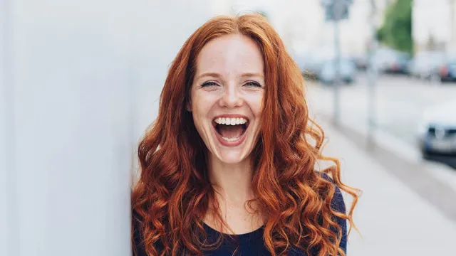 Smiling young woman with long curly red hair standing outdoors against a light wall, bright urban background.