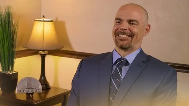 Smiling bald man in a suit sitting next to a lamp and potted plant on a wooden table with a welcome sign.