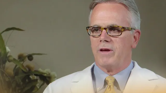 Middle-aged man in white coat and glasses speaking indoors with plants in the background.