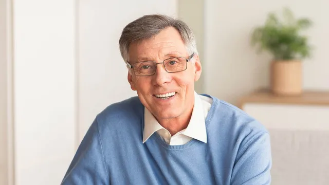 Smiling senior man with gray hair and glasses wearing a blue sweater in a bright, cozy room.