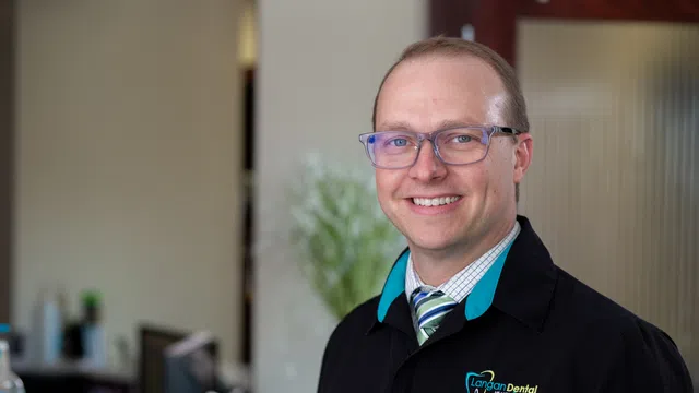 Smiling male dentist wearing glasses and Langan Dental jacket standing in dental office with blurred background.