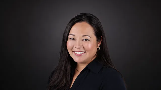 Smiling woman with long black hair wearing a dark blouse and silver hoop earrings against a black background