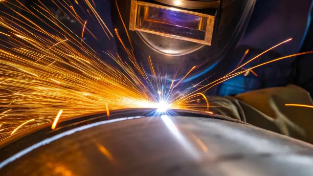 Welder wearing protective helmet sparks flying while welding a large metal pipe in industrial setting