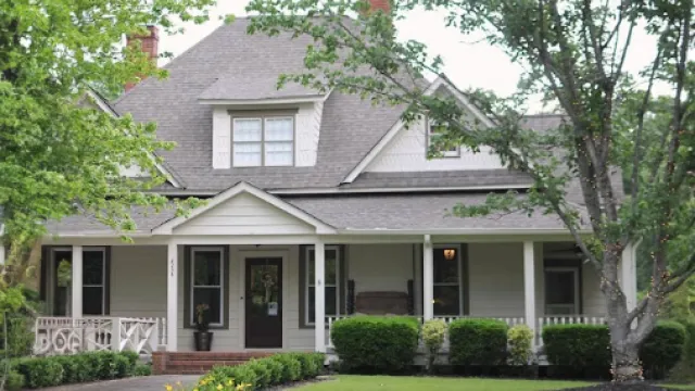 Charming suburban house with a wraparound porch and lush greenery in front.