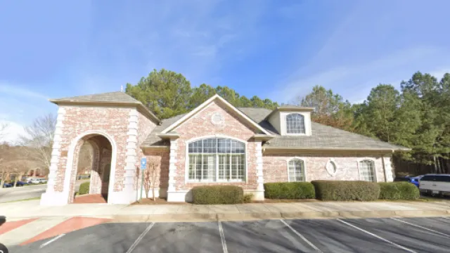 Brick building with large windows surrounded by trees and parked cars on a sunny day.