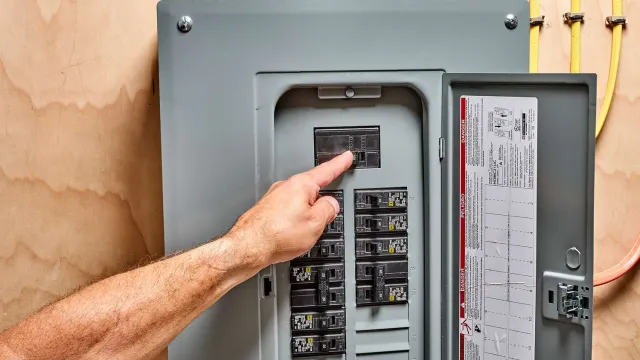 Hand flipping a circuit breaker switch in a gray electrical panel mounted on a wooden wall.