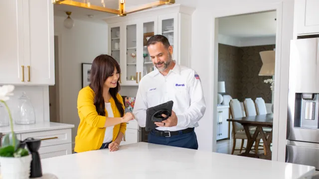 Technician in uniform showing a tablet to a woman in a modern white kitchen with stainless steel appliances.
