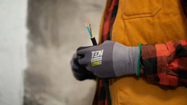 Electrician hand in protective glove holding stripped electrical wire indoors, wearing orange vest and plaid shirt.