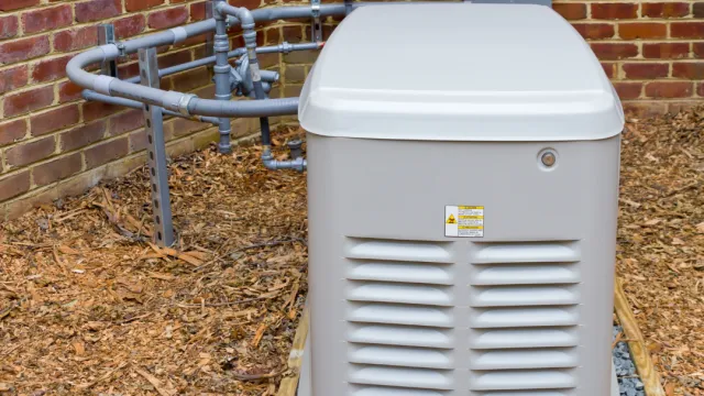 A grey generator installed outside a brick wall, surrounded by mulch and piping.