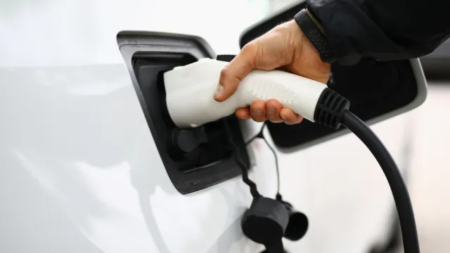 Person plugging a white electric vehicle charger into a white car at a charging station outdoors.