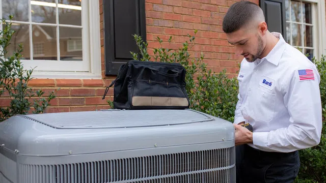 Technician inspecting an outdoor HVAC unit in front of a brick house, with a tool bag nearby.