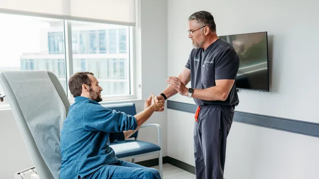 Physical therapist assisting man seated on exam table with wrist stretch in modern clinic room.