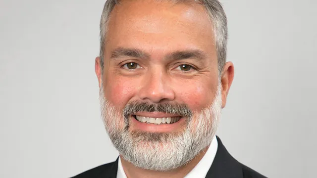 Professional portrait of a man in a suit with a beard, smiling against a neutral background.