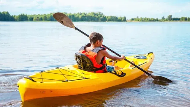 Young boy wearing life vest kayaking on calm water near tree-lined shore on a sunny day