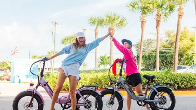 Two women high-fiving while standing next to electric bikes on a sunny street with palm trees.