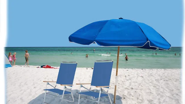 Two blue beach chairs under a blue umbrella on white sand with people swimming in the ocean on a sunny day.