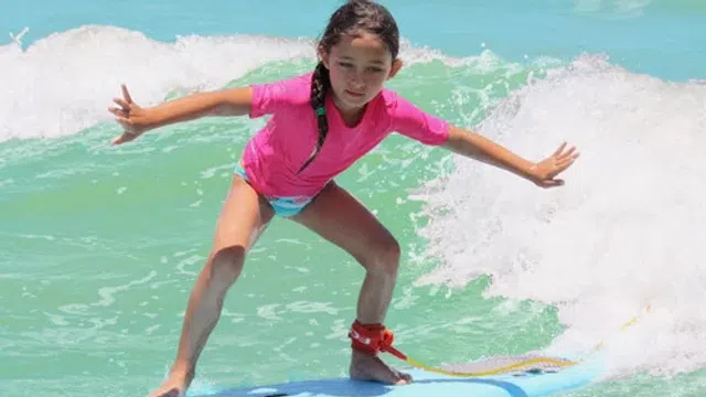 Young girl in a pink rash guard surfing on a blue board in clear ocean waves.