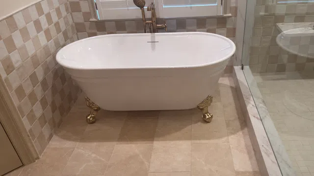 White clawfoot bathtub with gold accents next to a glass shower in a beige tiled bathroom.