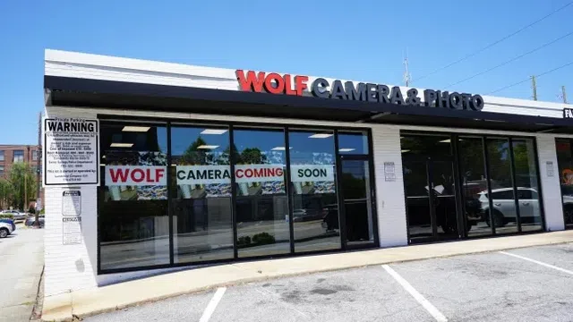 Wolf Camera & Photo store exterior with large signage and 'Coming Soon' banners on glass windows in a parking lot.
