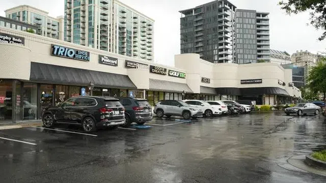 Shopping plaza with various stores and parked cars on wet pavement under overcast sky.