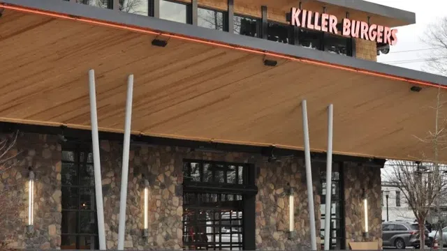 Killer Burgers restaurant exterior with stone wall, large windows, and wooden canopy supported by angled poles.