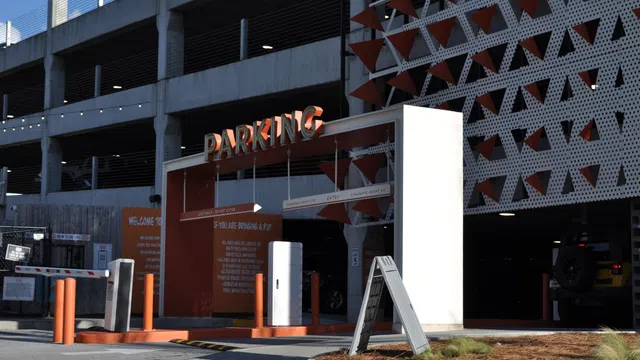 Entrance to a multi-level parking garage with orange and white signage and barriers under daylight.