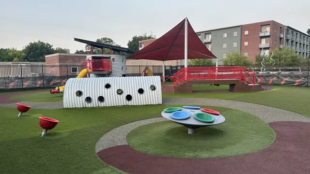 Modern outdoor playground with colorful equipment, climbing tunnel, shaded area, and rubber safety flooring under clear sky