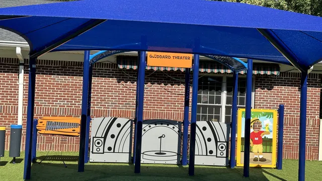 Outdoor children's play theater area with blue canopy, stage backdrop, and artificial grass seating resembling rocks.