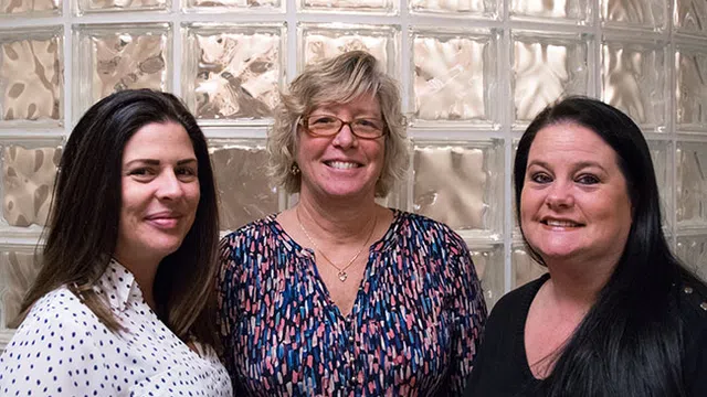 Three women smiling standing in front of a textured glass block wall, wearing casual patterned and solid tops.
